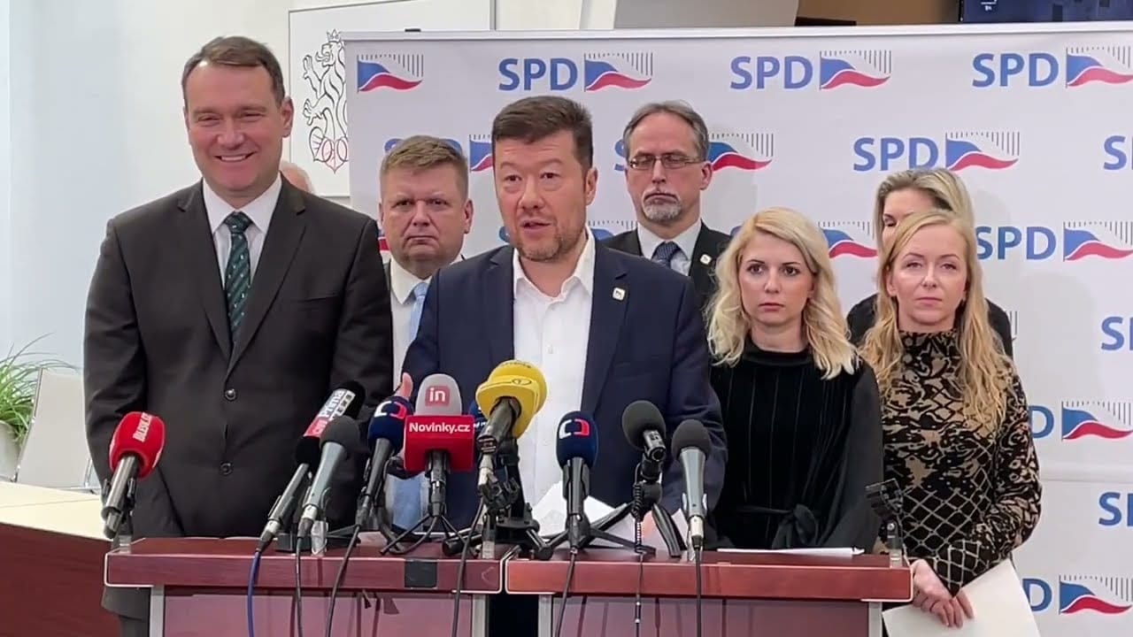 Tomio Okamura speaking at a lectern with microphones in front of a backdrop with SPD logos, surrounded by six other people
