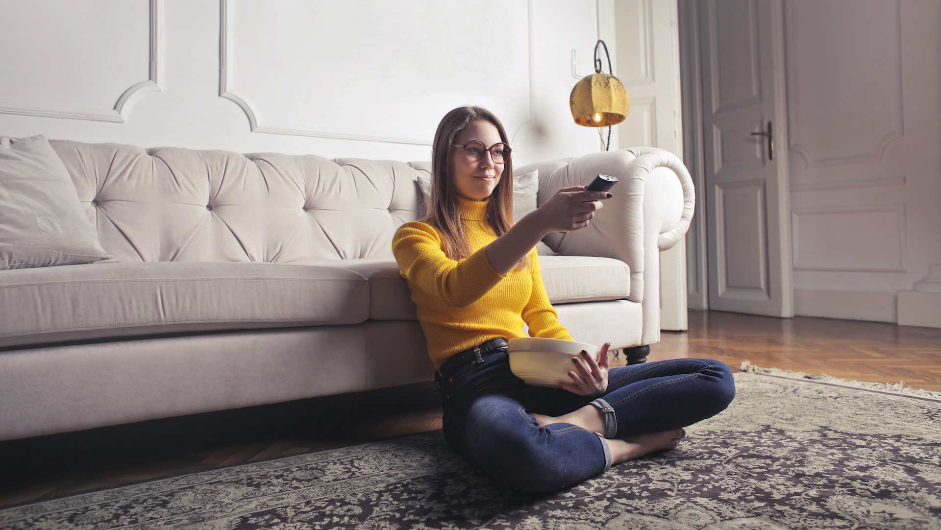 Woman in a yellow sweater sitting on the floor by a sofa, pointing a TV remote and holding a bowl