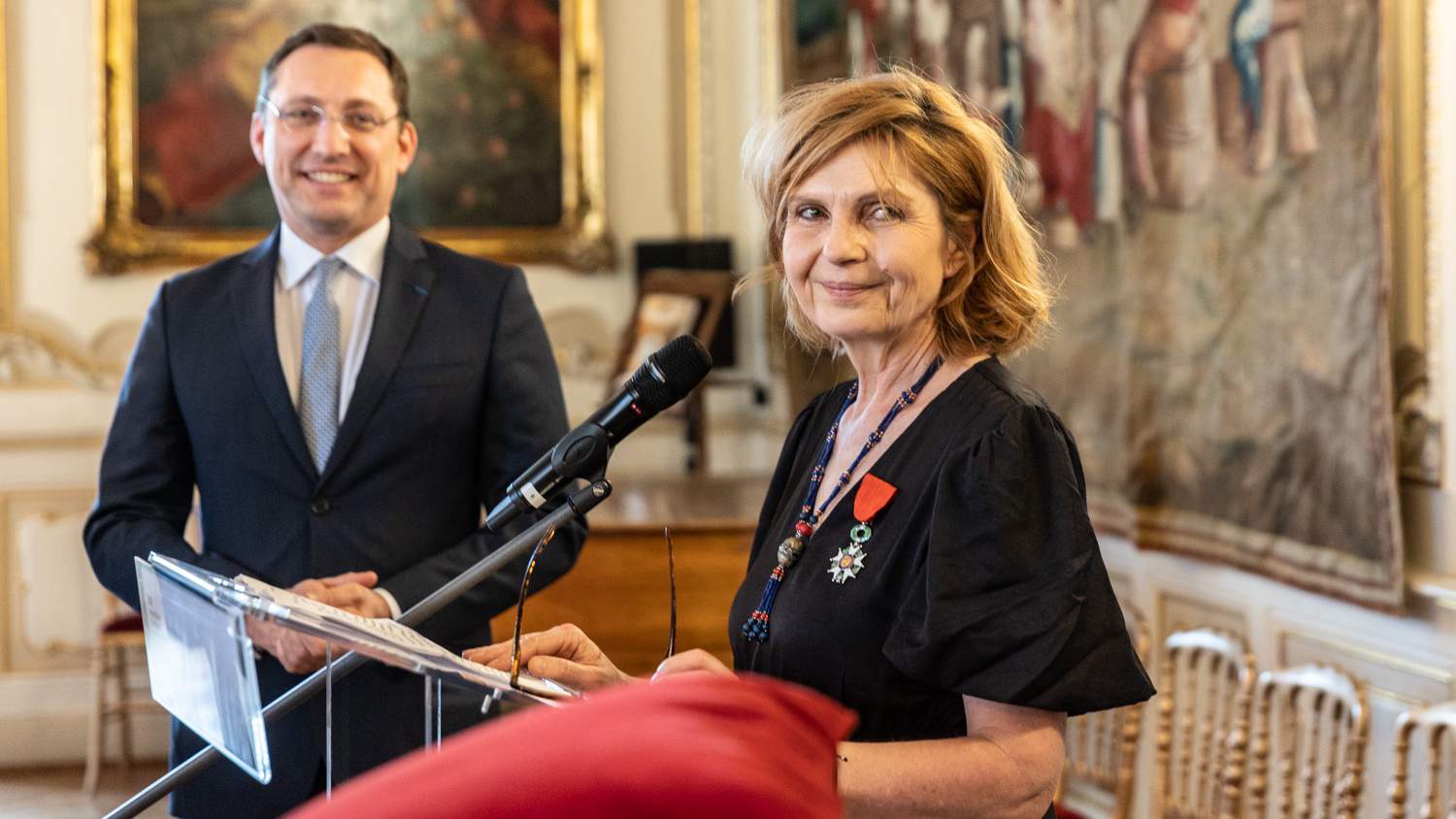 Journalist Petra Procházková in a black dress with a red-ribboned medal standing at a lectern with a microphone, a smiling man in a dark suit behind her in an ornate hall