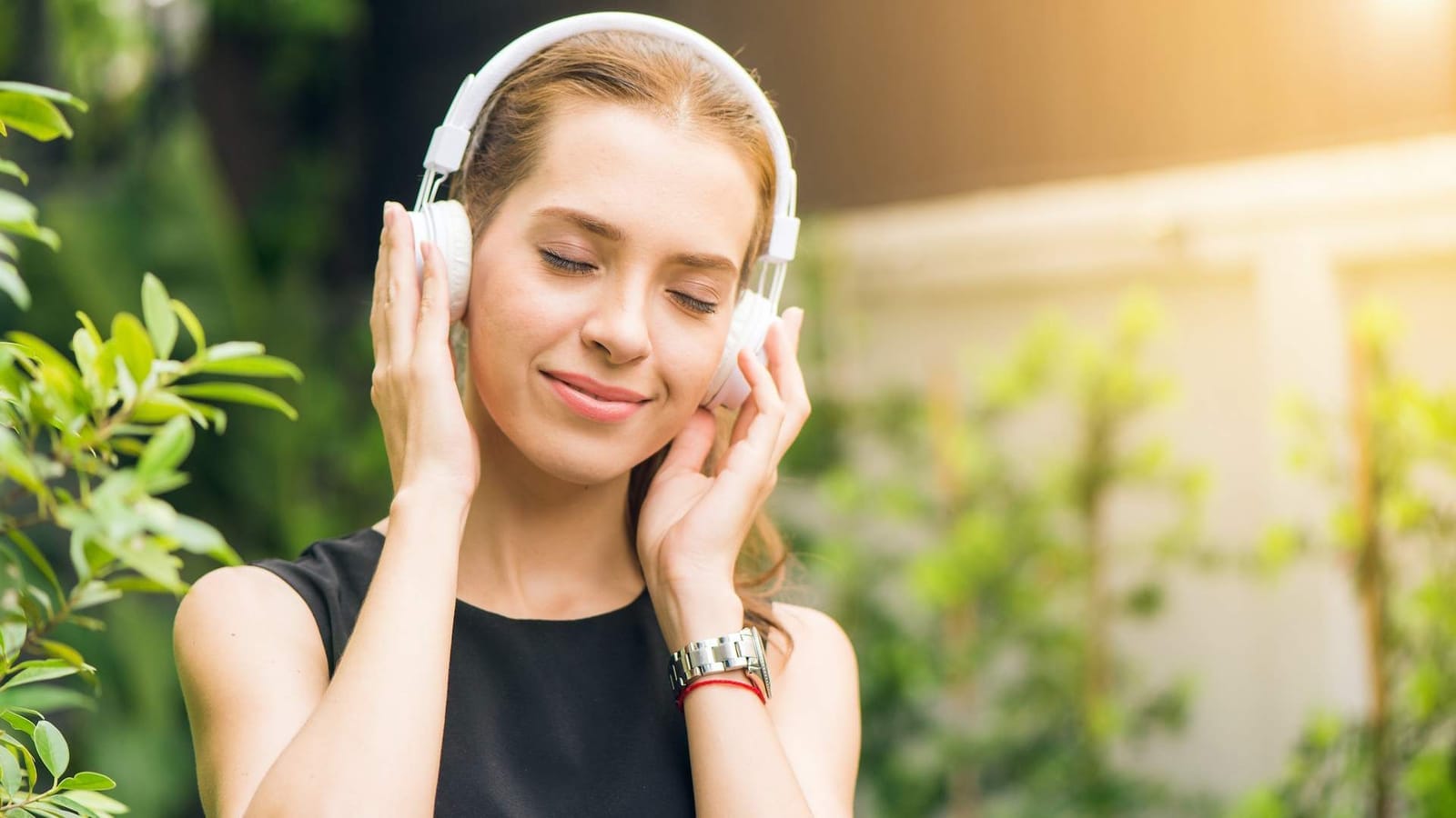 Smiling young woman with closed eyes holding white over-ear headphones, standing outdoors among green plants