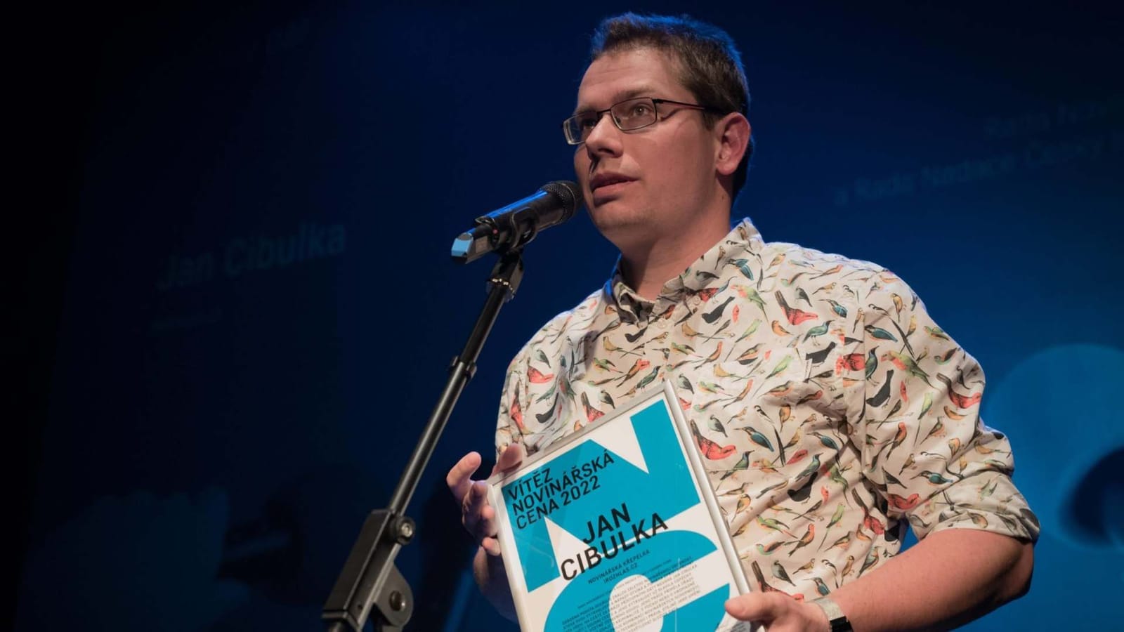 Jan Cibulka speaking into a microphone on stage, holding a certificate reading "Vítěz Novinářská cena 2022, Jan Cibulka"