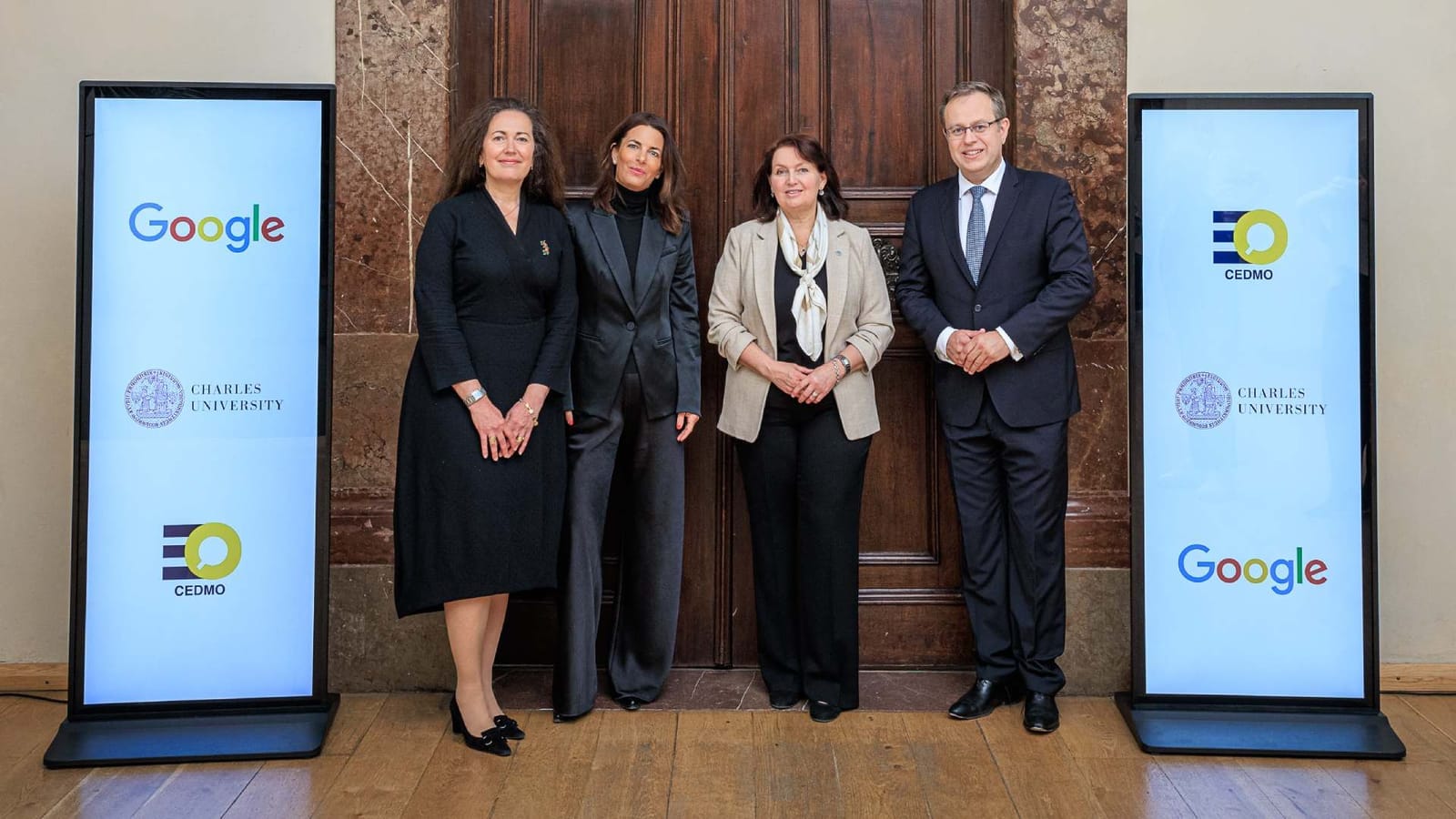 Three women and a man stand between two digital displays showing the Google, Charles University and CEDMO logos