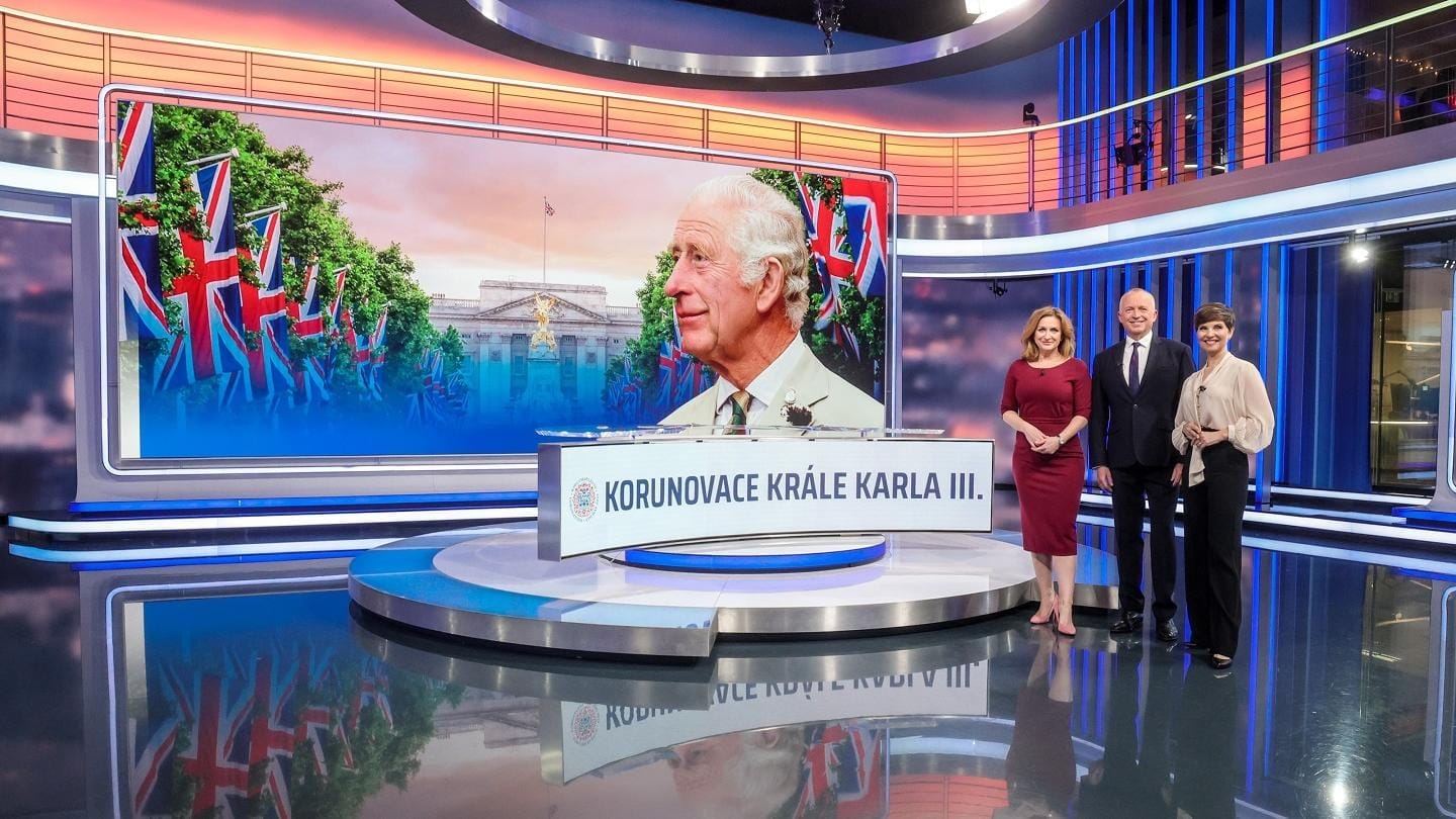 TV studio with a large screen showing King Charles III and the caption "Korunovace krále Karla III.", three presenters standing beside a desk