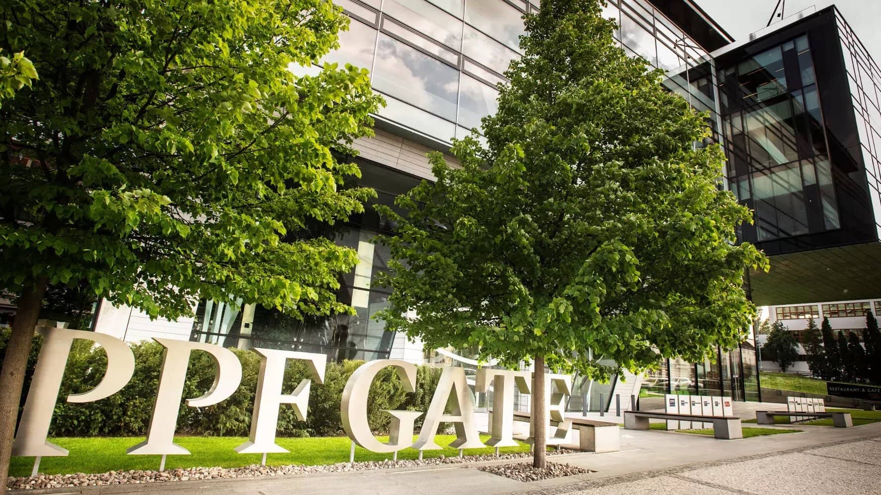 Large white "PPF GATE" lettering in front of a glass office building, with trees in the foreground