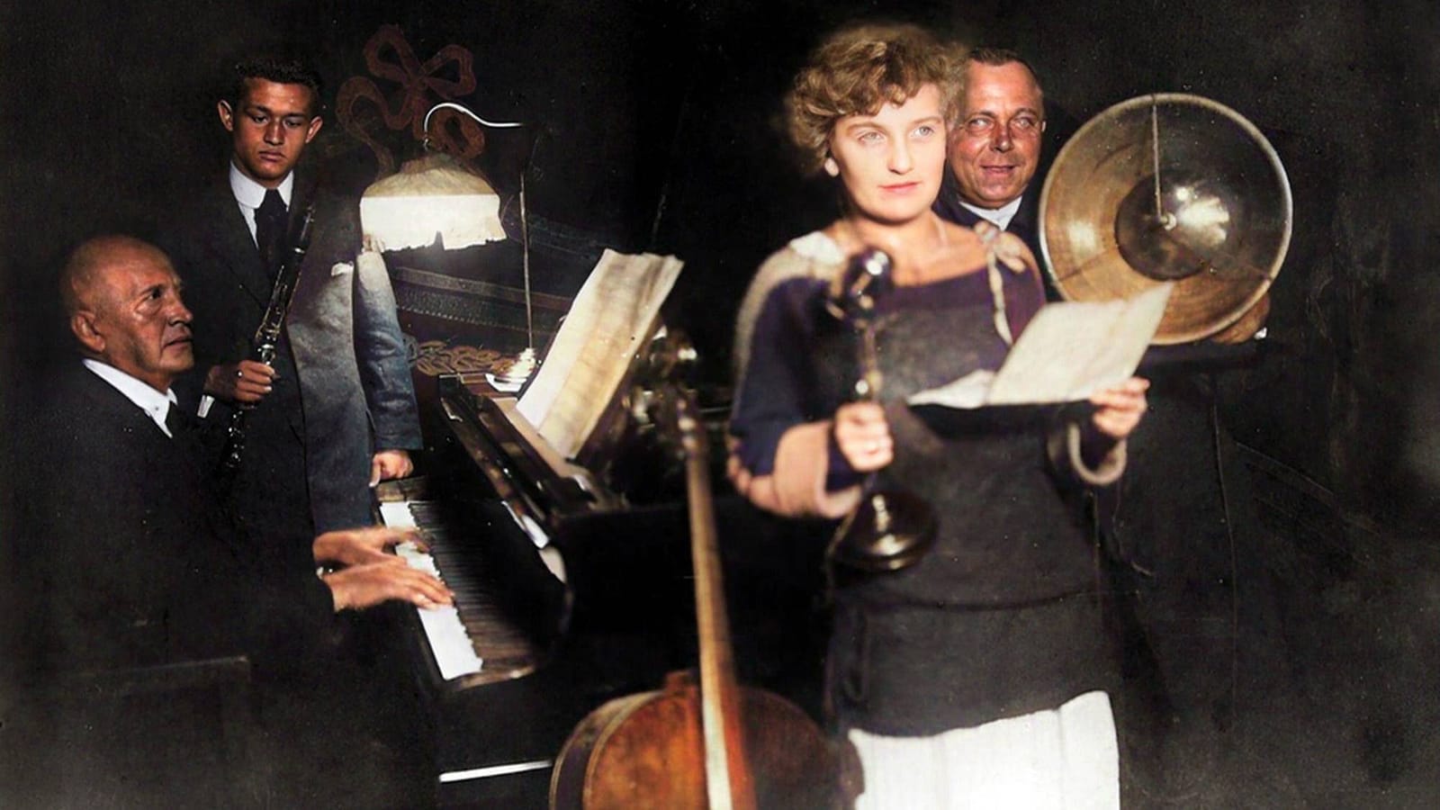 Colourised historical photo of an early radio studio: a woman reads from a sheet into a microphone, a man plays piano, another holds a clarinet, with a large horn loudspeaker behind