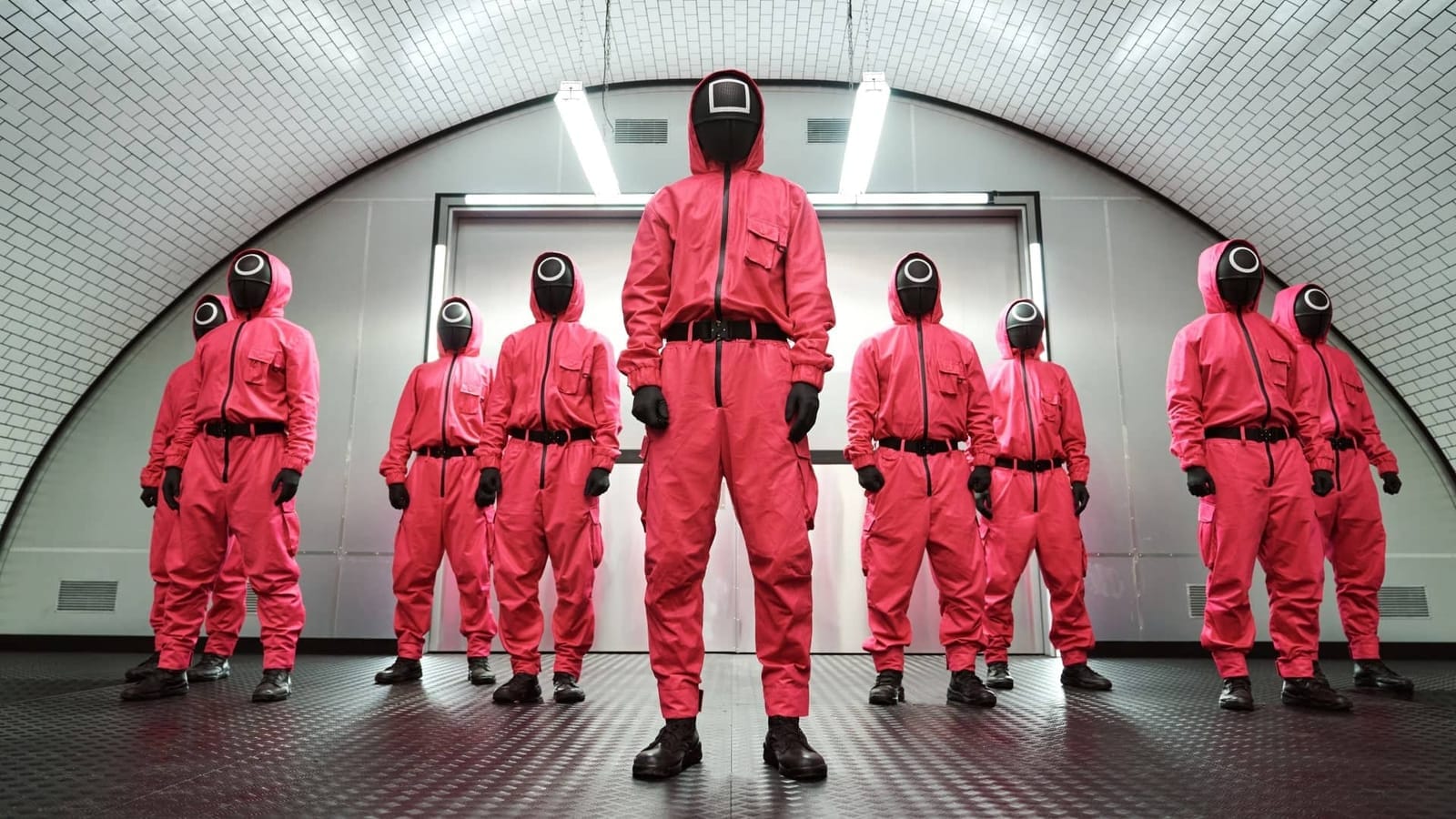 Guards in pink jumpsuits and black masks with geometric symbols standing in a tiled corridor, a scene from Squid Game